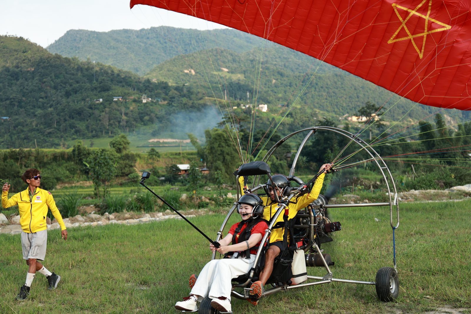 Khách ngồi trên paramotor đôi cười tươi, phi công phía sau, cánh dù đỏ sao vàng đang dựng lên trước khi cất cánh